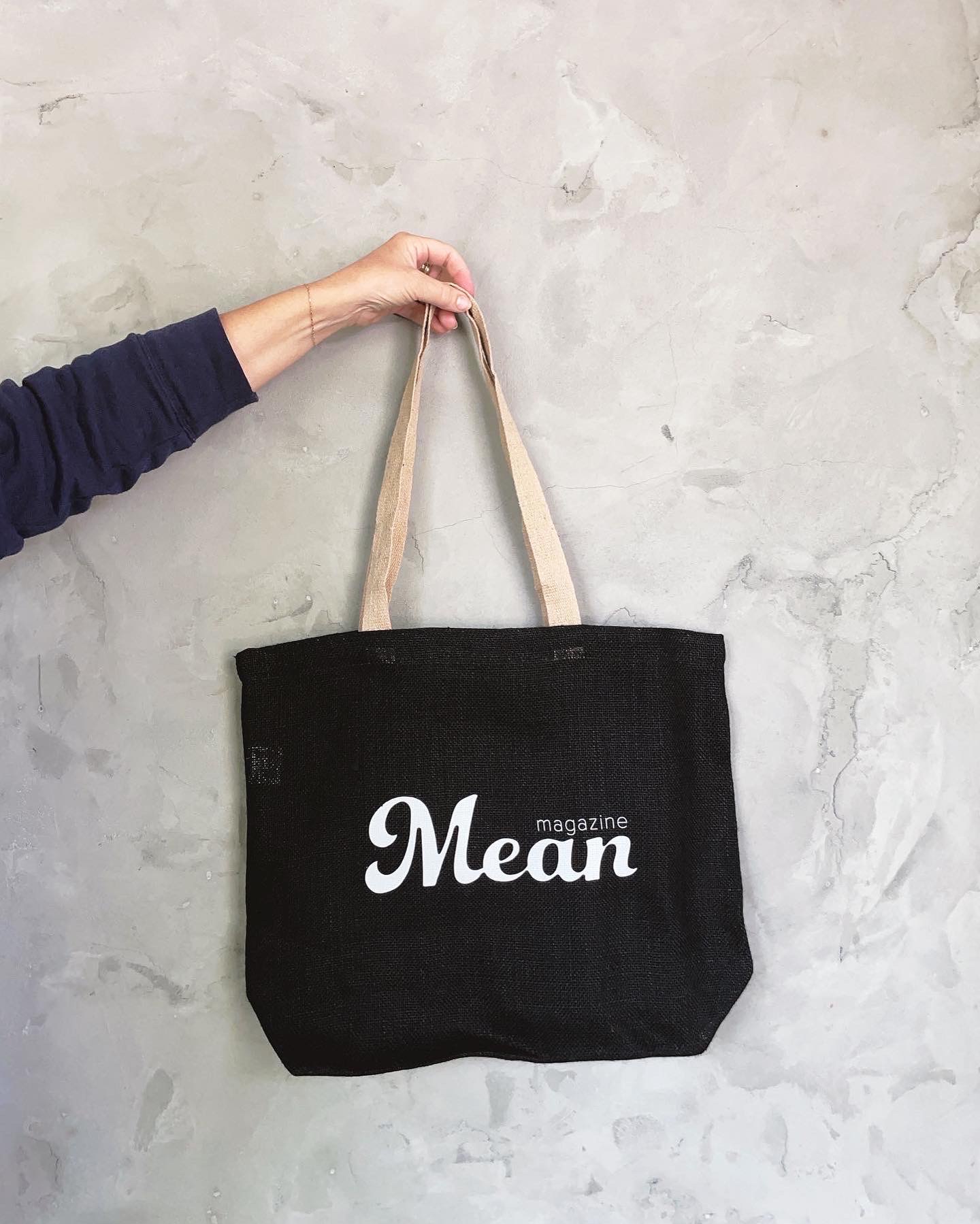 a disembodied arm holds a black mean magazine jute tote bag